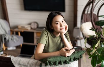 A young woman in a green t-shirt sits indoors, leaning on a cushion, holding a smartphone, and looking at the camera. A laptop and home decor are visible in the background.