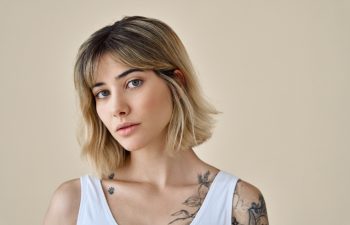 Cool blonde pretty gen z girl female stylish model short blonde hair beautiful face healthy skin and tattoos looking at camera wearing white top isolated at beige background. Portrait. Copy space